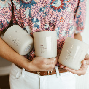 Spring and summer candles in cream vessels surrounded by lilac trimmings, watermelon slice, and cucumber.